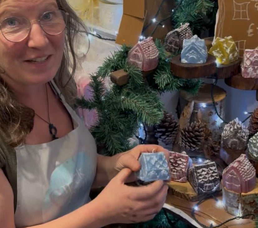 A woman wearing glasses and a white apron smiles while holding a small decorated soap. She is standing in front of a decorated display of handmade soaps, pinecones, and a chalkboard sign in a cozy indoor setting.
