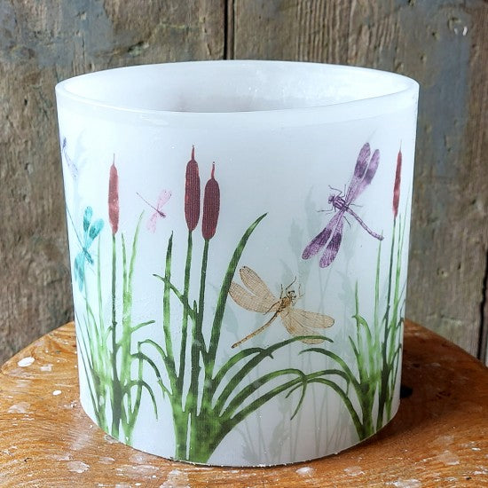 White wax hurricane candle  with nature-themed design featuring plants and dragonflies on a wooden surface.