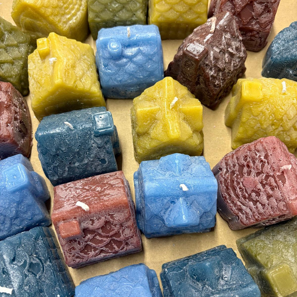A collection of handcrafted, cube-shaped beeswax candles in various colors including blue, yellow, green, and red, arranged on a tan surface.