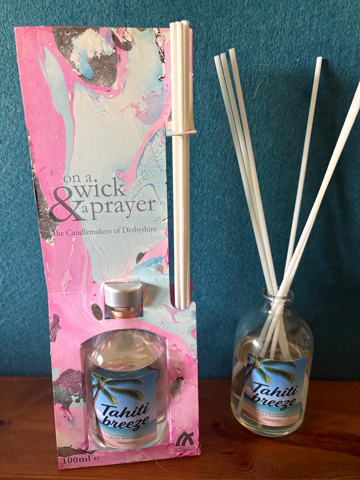 Tahiti Breeze reed diffuser set by on a. wick & prayer, featuring a pink and blue branded cardboard box with a glass bottle and several white reeds, displayed on a wooden surface against a dark blue wall.