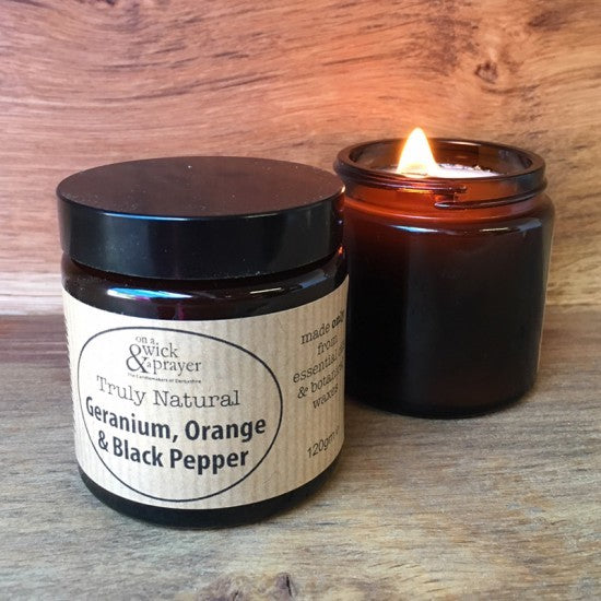  'Truly Natural Geranium, Orange & Black Pepper' candle with a lit candle on a wooden surface