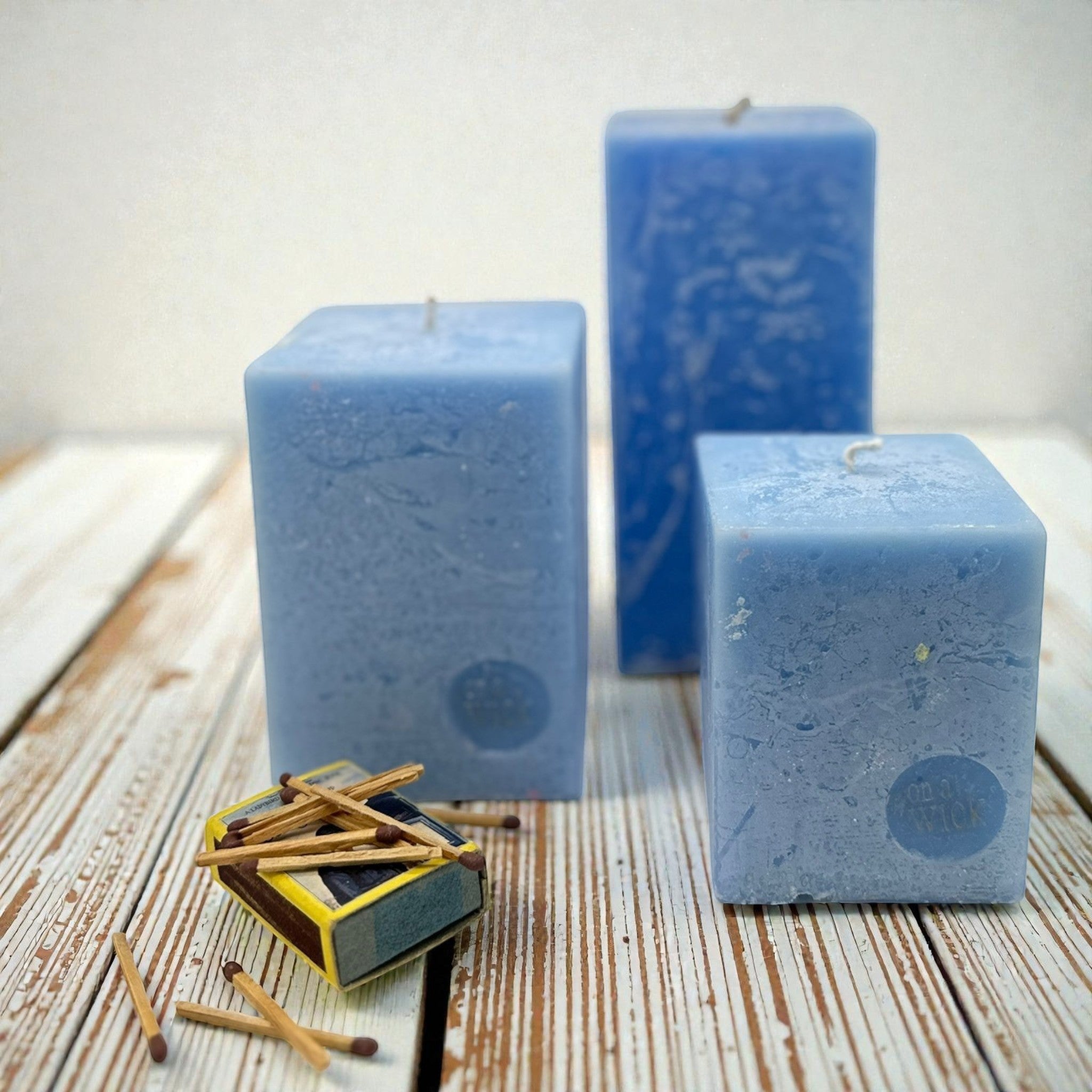 On a Wick and a Prayer blue scented square pillar candles in varying sizes, with a matchbox and scattered matches.