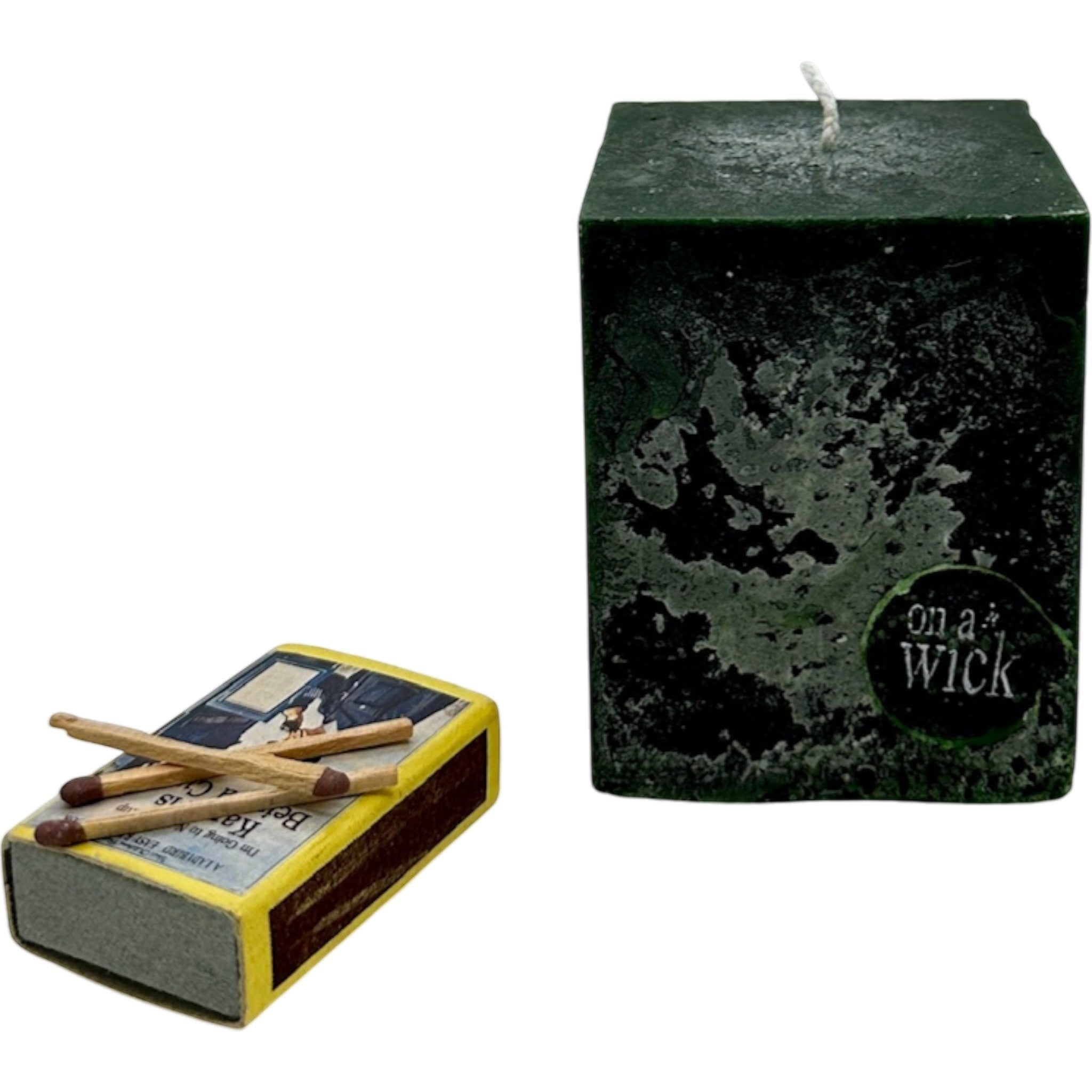 On a Wick and a Prayer dark green square pillar candle with a textured finish and a white wick, next to a vintage matchbox.