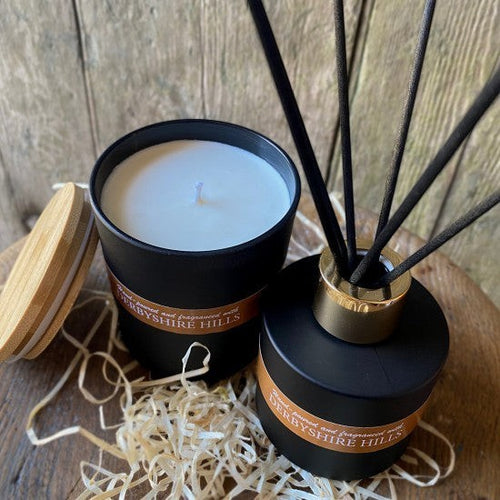 On a Wick and a Prayer Derbyshire Hills candle and reed diffuser set in black jars with gold accents, featuring a white candle and black reeds.