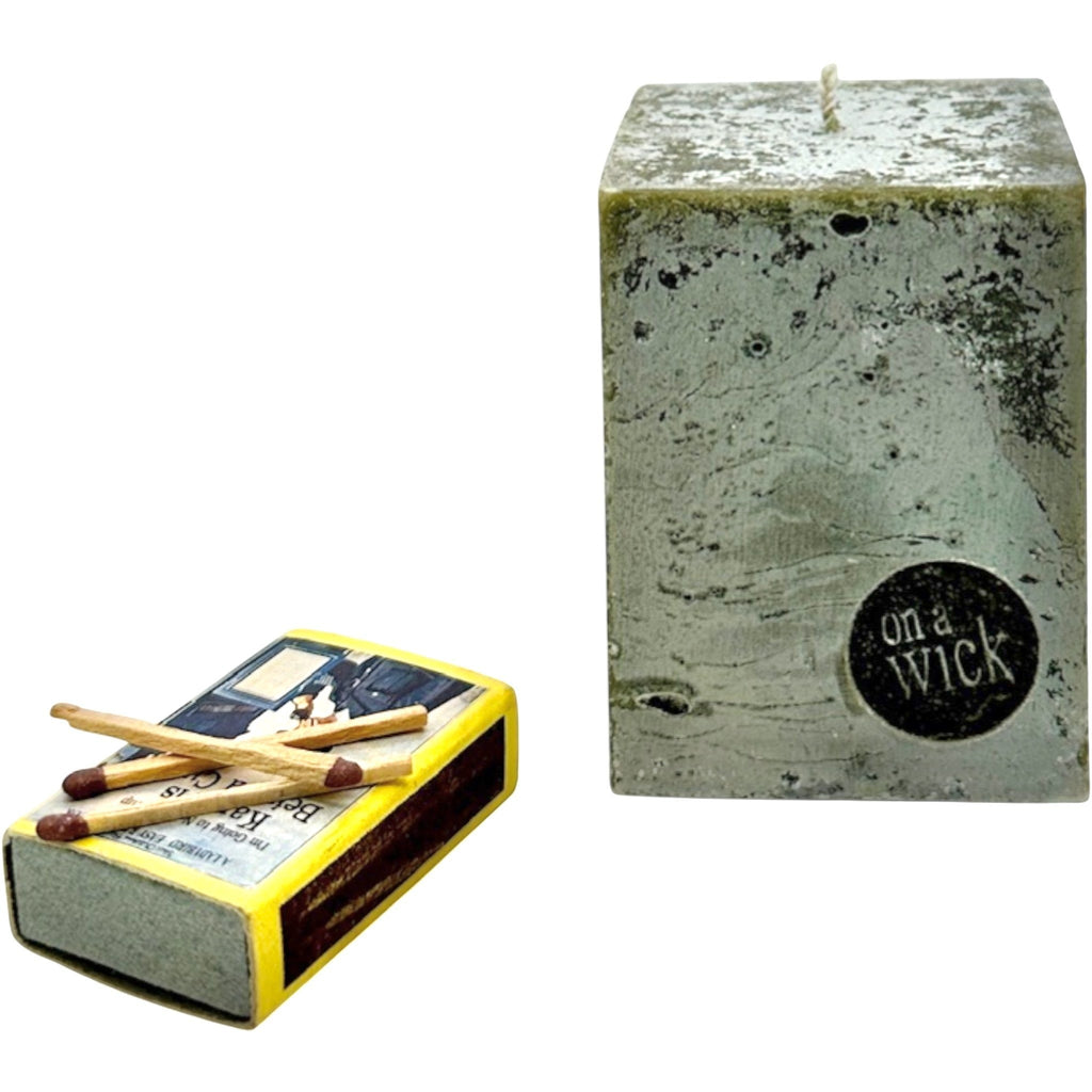 On a Wick and a Prayer grey textured square pillar candle with a wick and a black circular logo. A vintage matchbox with matches rests beside it.