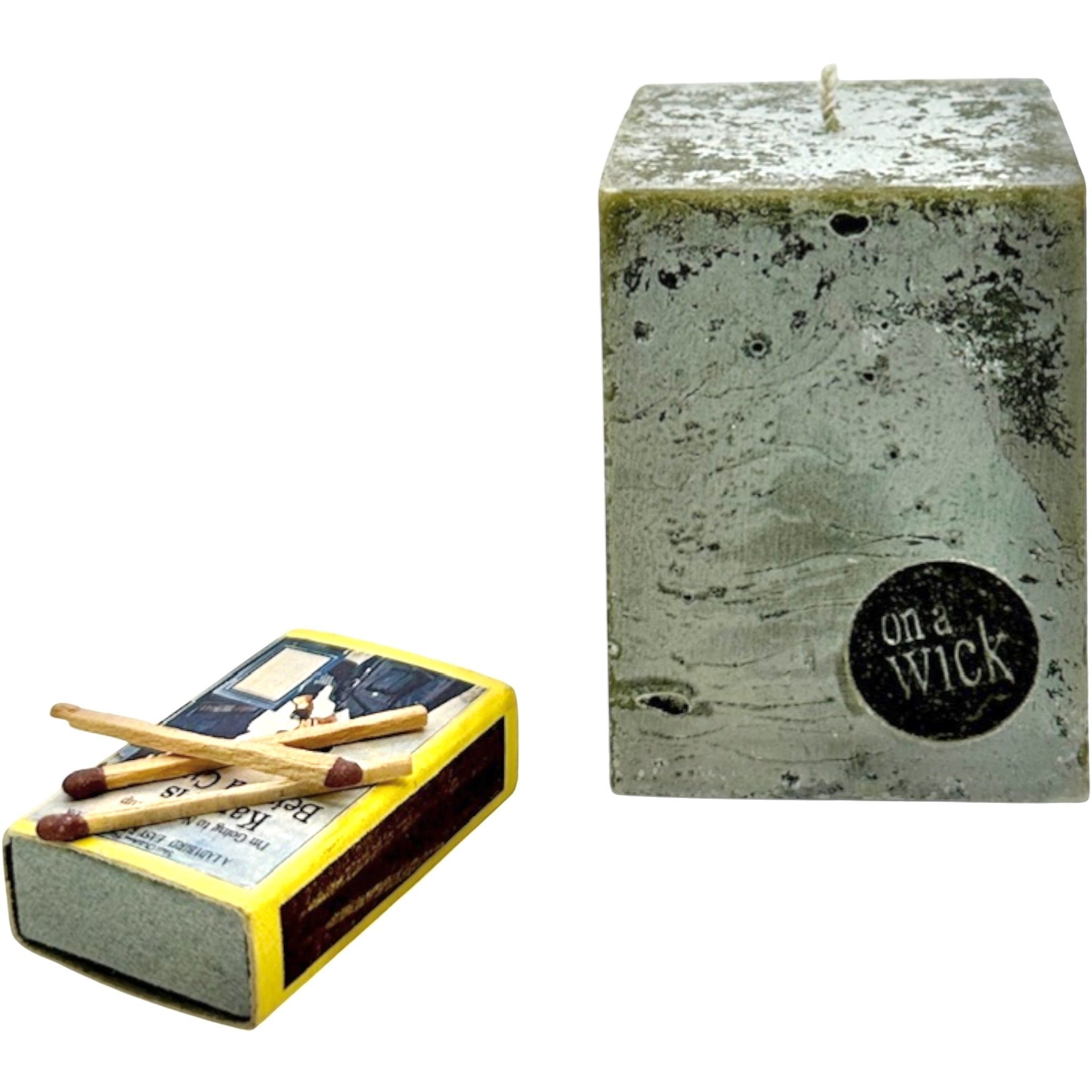 On a Wick and a Prayer grey textured square pillar candle with a wick and a black circular logo. A vintage matchbox with matches rests beside it.