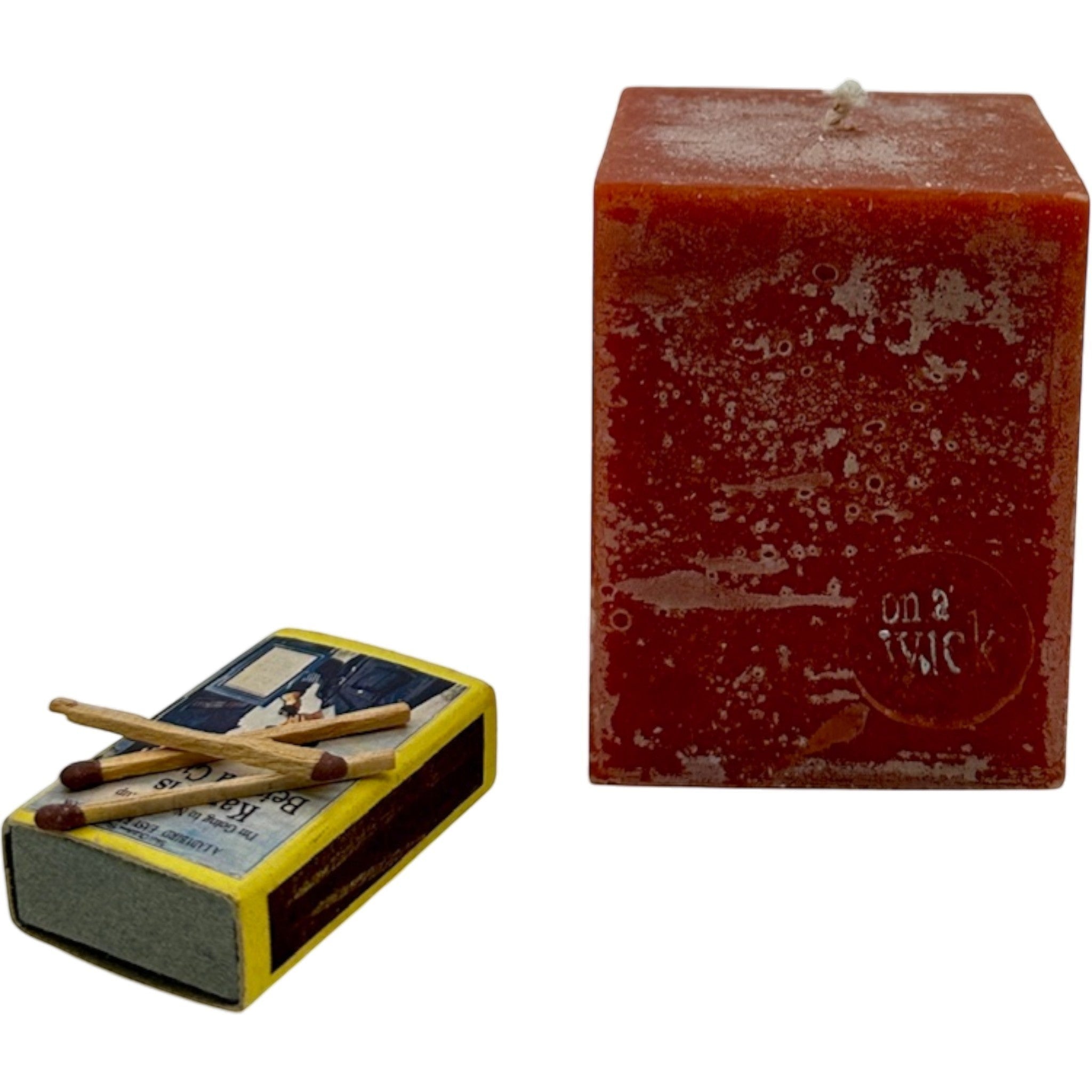 On a Wick and a Prayer red square pillar candle with a rustic finish, next to a vintage matchbox.