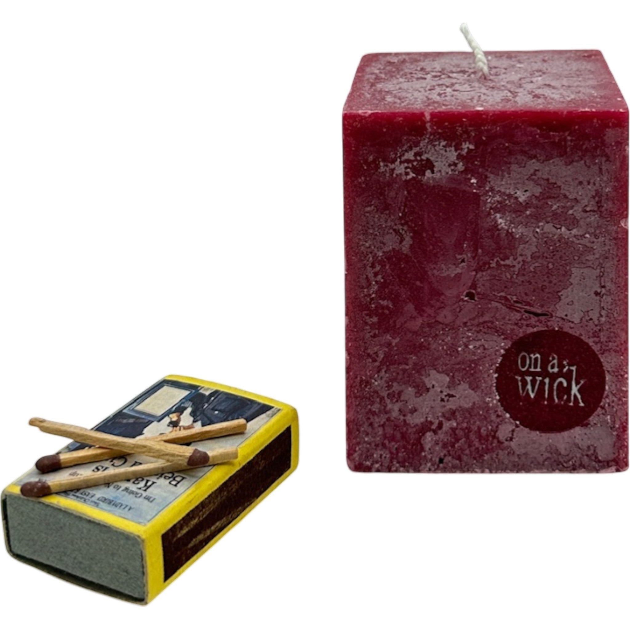 On a Wick and a Prayer deep red square pillar candle with a rustic finish, next to a vintage matchbox.