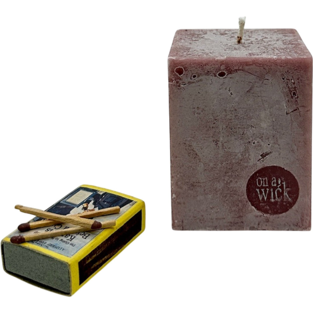 On a Wick and a Prayer rustic red square pillar candle with a textured finish, next to a vintage-style matchbox.