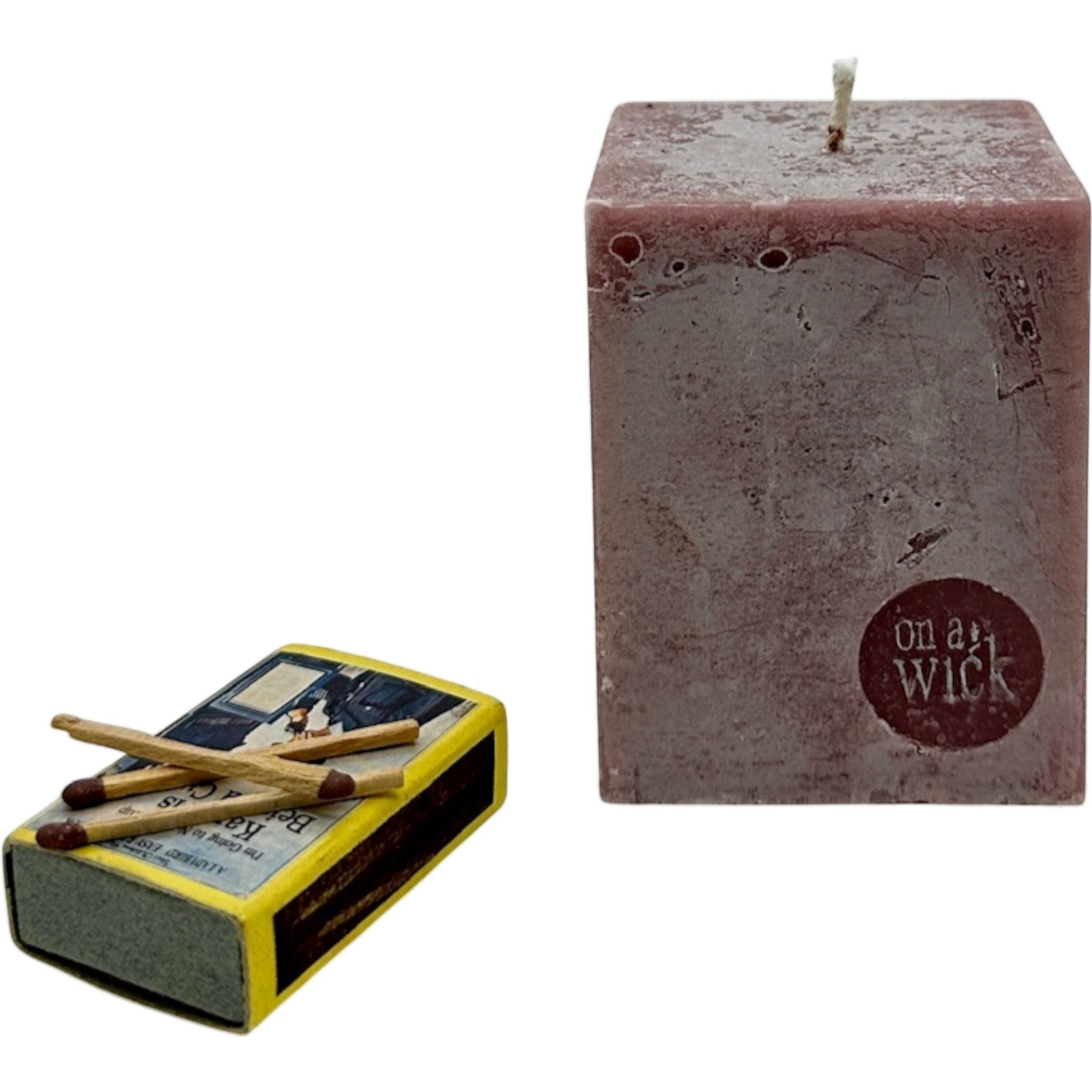 On a Wick and a Prayer rustic red square pillar candle with a textured finish, next to a vintage-style matchbox.